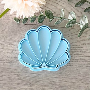 May include: A light blue plastic seashell cookie cutter. The cutter has a detailed design with multiple ridges.