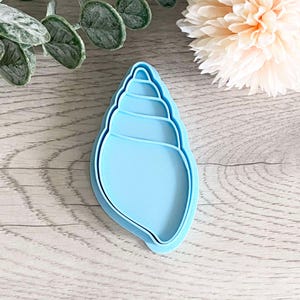 May include: A light blue plastic seashell cookie cutter. The cutter is shaped like a seashell with multiple layers.