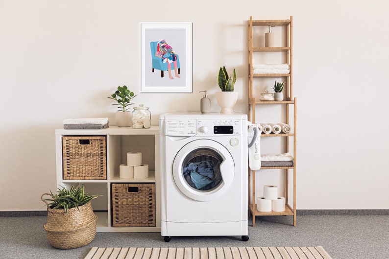 May include: A white washing machine with a glass door and a digital display. The washing machine is in a laundry room with a white shelf unit, a bamboo shelf unit, and a framed print of a person sitting in a chair.