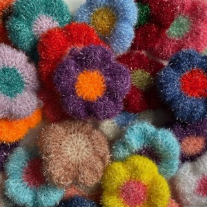 Può includere: Una collezione di spugne a forma di fiore fatte a mano. Le spugne sono realizzate con un filato strutturato e presentano una varietà di colori, tra cui rosso, viola, blu, verde, giallo e arancione. Le spugne hanno un diametro di circa 10 cm.