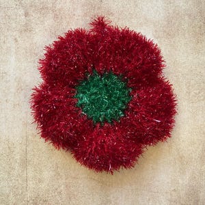 Può includere: Un fiore decorativo rosso e verde, realizzato con un materiale soffice. Il fiore è rotondo con un centro verde e petali rossi. Lo sfondo è di un colore neutro e strutturato.