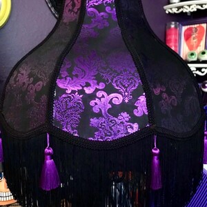 Pu&ograve; includere: Un paralume nero con un motivo floreale viola e frange nere. Il paralume ha un bordo decorativo e nappe viola. In stile gotico o vintage.