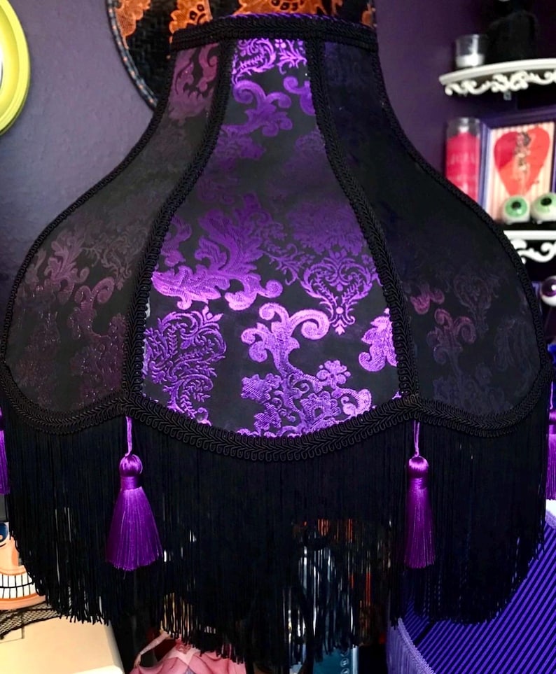 Pu&ograve; includere: Un paralume nero con un motivo floreale viola e frange nere. Il paralume ha un bordo decorativo e nappe viola. In stile gotico o vintage.