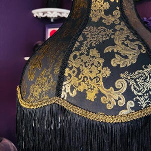 Pu&ograve; includere: Paralume nero con motivo floreale dorato e frange. Il paralume ha un bordo dorato e frange nere sul fondo. Un elemento di illuminazione decorativo.