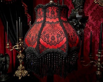 Könnte beinhalten: Ein roter und schwarzer Lampenschirm im gotischen Stil. Der Schirm hat eine rote Stoffbasis, die mit komplizierten schwarzen Spitzendesigns überzogen ist. Schwarze Perlenfransen hängen vom unteren Rand, was einen dekorativen Akzent setzt. Der Lampenschirm ist an einer Metallhalterung aufgehängt.
