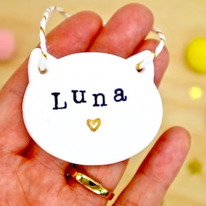 Personalised cat name hanging keepsake decoration. Cat shaped and made from white clay, with tiny gold love heart. Cat's name is stamped in black on the front. Finished with white and gold twine. Pet memorial gift or simply just because.