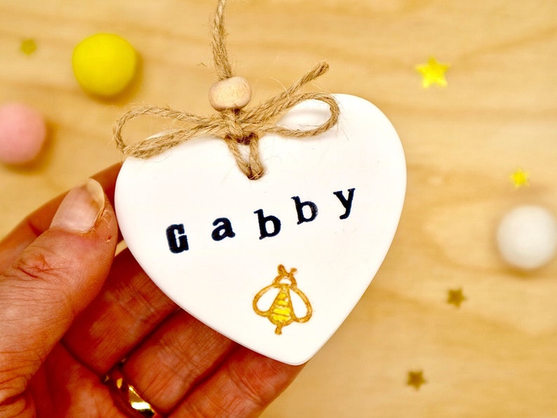 Personalised bee keepsake decoration, handmade from white clay, with a gold and yellow stamped bee. A hanging heart for birthdays or any other occasion. Tied with natural jute twine and wooden bead. Gift for friend, daughter, girl, mum.