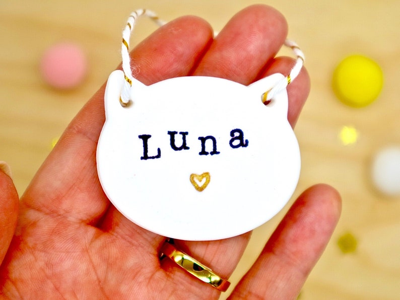 Personalised cat name hanging keepsake decoration. Cat shaped and made from white clay, with tiny gold love heart. Cat's name is stamped in black on the front. Finished with white and gold twine. Pet memorial gift or simply just because.