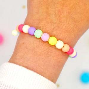 May include: A colourful beaded bracelet featuring round beads in pastel shades of pink, yellow, green, purple, and white. The bracelet is worn on a wrist, highlighting the vibrant accessory. A playful piece of jewellery.