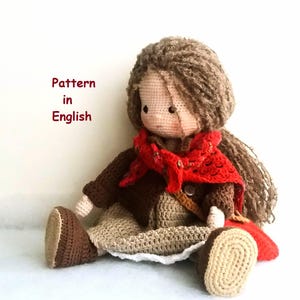 Può includere: Bambola all'uncinetto con capelli castani, vestita con un abito rosso e marrone. La bambola è seduta su una superficie bianca. Il testo "Pattern in English" è visibile nell'immagine.