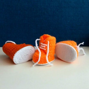 May include: Three crocheted baby booties in orange and white. The booties have a lace-up design and are made with a soft, textured yarn.