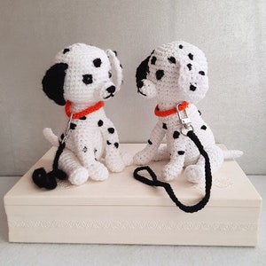 Può includere: Due peluche di cane dalmata all'uncinetto con macchie nere e collari arancioni. I peluche sono seduti su una scatola bianca con un guinzaglio nero.