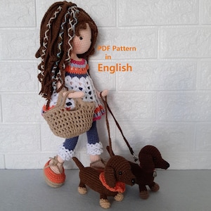 May include: A handmade crochet doll with brown hair and a white dress, carrying a basket and walking two brown dachshund dogs on leashes. The doll is wearing orange shoes. Text on the image reads "PDF Pattern in English."