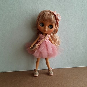 May include: A pink and white crocheted dress with a matching crocheted bag and headband. The doll has blonde hair and blue eyes.