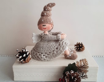 May include: A crocheted doll wearing a brown and white hat and a beige dress. The doll is sitting on a white box with a pine cone and greenery in front of it.