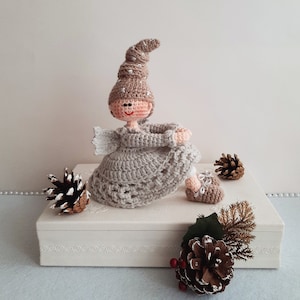 May include: A crocheted doll wearing a brown and white hat and a beige dress. The doll is sitting on a white box with a pine cone and greenery in front of it.
