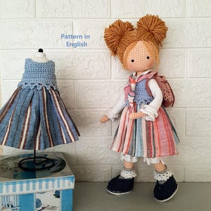 以下が含まれることがあります： 手作りのかぎ針編みの人形。糸の髪、ストライプのドレス、青いベスト、ネイビーの靴を着用。お揃いのドレスがスタンドに展示されています。「Pattern in English」の文字が見えます。