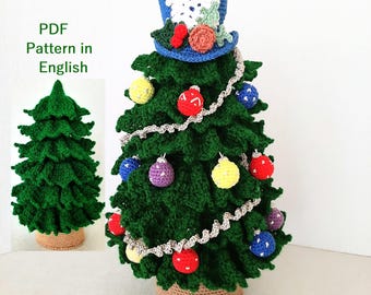 Op de afbeelding: Een handgemaakte groene gehaakte kerstboom versierd met kleurrijke ornamenten en een blauwe hoge hoed. De boom is versierd met rode, gele, paarse en blauwe ornamenten. De boom staat in een geweven mand. De tekst "PDF Pattern in English" staat in de afbeelding.