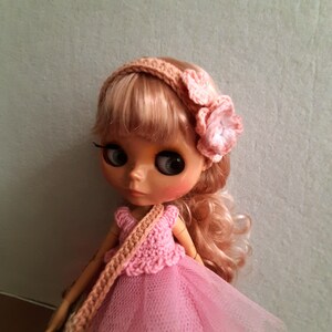 May include: A doll with blonde hair wearing a pink crocheted dress and a headband with pink flowers.