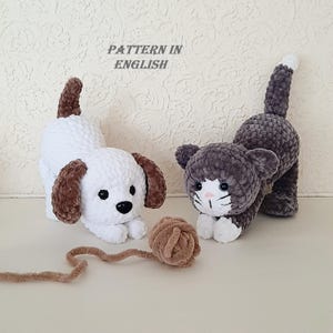 Pode incluir: Dois brinquedos de crochê de pelúcia, um cão branco com orelhas castanhas e um gato cinzento e branco, ambos numa pose brincalhona. Um pequeno novelo de lã bege encontra-se entre eles. O texto "PATTERN IN ENGLISH" é visível acima dos brinquedos.