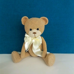 Może przedstawiać: Pluszowy miś w kolorze brązowym, wykonany na szydełku, z kremową satynową kokardką. Miś siedzi na białym tle.