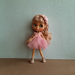 May include: A pink and white crocheted dress with a tulle skirt and a flower headband. The doll has blonde hair and blue eyes.