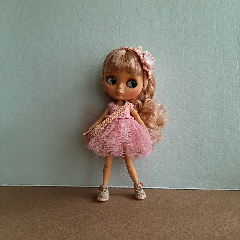 May include: A pink and white crocheted dress with a tulle skirt and a flower headband. The doll has blonde hair and blue eyes.
