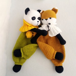 May include: Two crocheted stuffed animals, a panda and a fox, are hugging. The panda is wearing a yellow and green outfit and the fox is wearing a brown and white outfit.