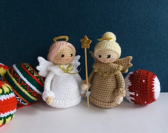 May include: Two crocheted angels, one in white with a pink face and one in brown with a yellow face, stand on a white surface with a star-shaped wand between them.  There are three red and green crocheted ornaments in the background.