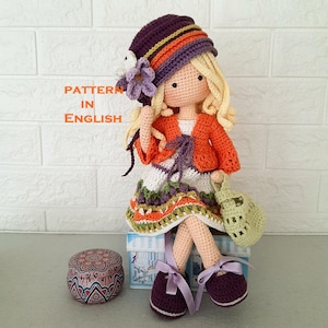May include: A handmade crochet doll with blonde hair, wearing a purple hat, orange jacket, and a white and green dress. The doll is holding a small green bag and is sitting on a box. The text "PATTERN IN ENGLISH" is visible.