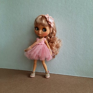 May include: A pink and white crocheted dress with a tulle skirt and a flower headband on a doll with brown hair and brown eyes.