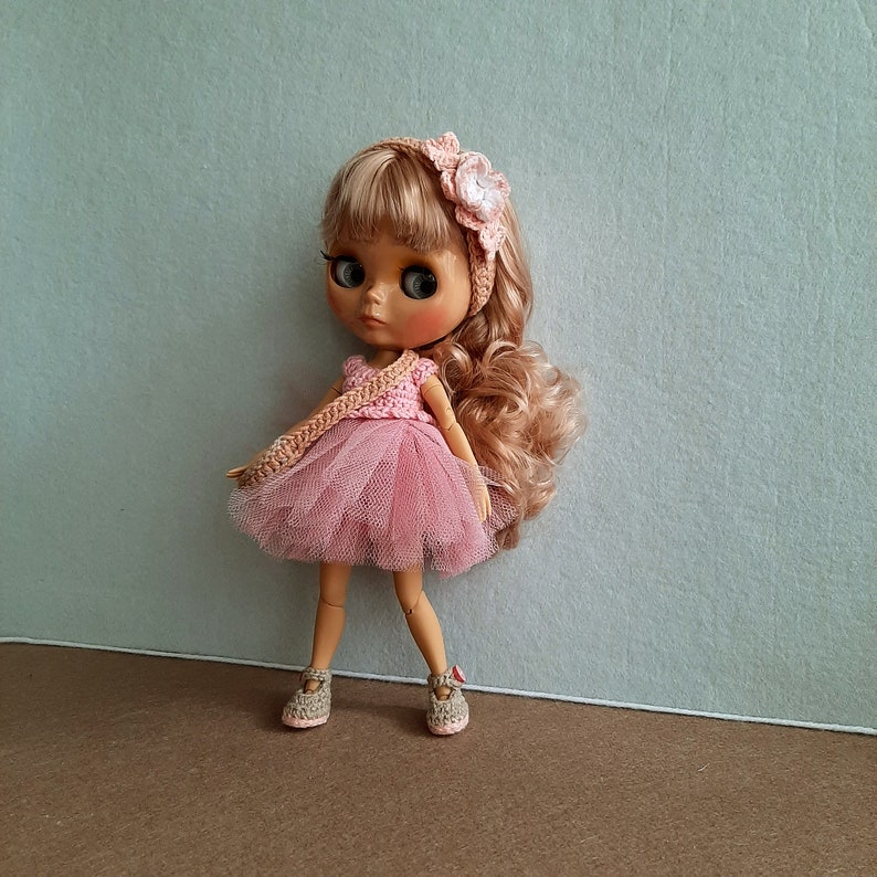 May include: A pink and white crocheted dress with a tulle skirt and a flower headband on a doll with brown hair and brown eyes.