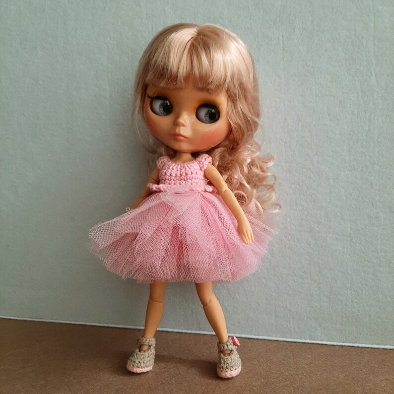 May include: A doll with blonde hair wearing a pink tulle dress and crocheted bodice. The doll is wearing brown shoes.