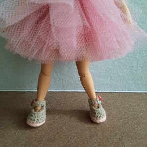 May include: A doll wearing a pink tulle skirt and brown crocheted shoes with red accents.