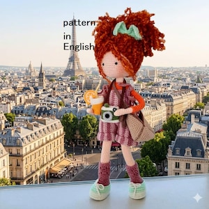 May include: A handmade crochet doll with red hair, a green bow, and a pink dress, holding a camera and a drink. The doll is wearing a shoulder bag and standing in front of a cityscape with the Eiffel Tower in the background. The text "pattern in English" is visible.