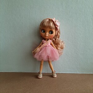 May include: A pink and white crocheted dress with a tulle skirt and a flower headband. The doll has blonde hair and blue eyes.