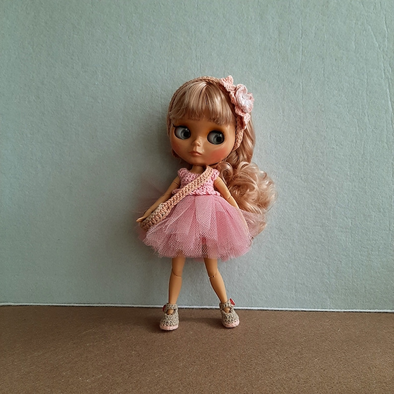 May include: A pink and white crocheted dress with a tulle skirt and a flower headband. The doll has blonde hair and blue eyes.