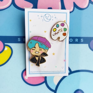 May include: Two enamel pins featuring a cartoon character with blue hair and a purple beret and a white paint palette with the word "VANTE" written on it.