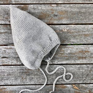 Könnte beinhalten: Eine graue gestrickte Baby-Mütze mit einer langen Bindekordel. Die Mütze liegt auf einer Holzoberfläche.