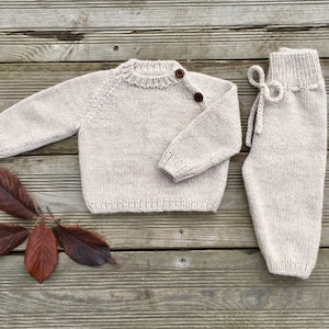 Op de afbeelding: Een gebreide babytrui en broekset in een licht beige kleur. De trui heeft een ronde hals en lange mouwen. De broek heeft een trekkoord in de taille.