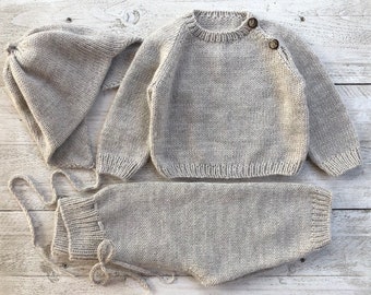 Op de afbeelding: Een grijze gebreide baby-outfit met een trui, broek en een muts. De trui heeft een ronde hals en knoopsluitingen op de schouder.