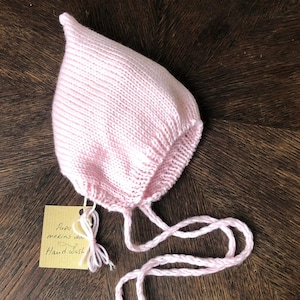 May include: A pink knitted baby bonnet with ties. The tag reads "Fine merino wool Hand wash."