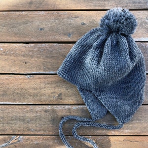 Puede incluir: Un gorro de punto gris con un pompón esponjoso en la parte superior. El gorro tiene orejeras y una larga cuerda de atar.