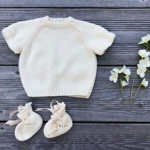Puede incluir: Un jersey de punto blanco para beb&eacute; con mangas cortas y cierre de botones. El jersey est&aacute; colocado sobre una superficie de madera con flores blancas.