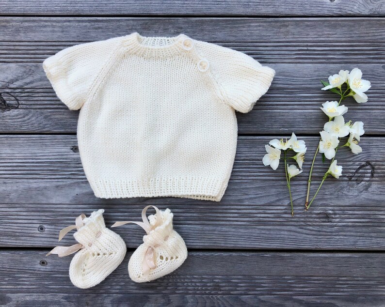 Puede incluir: Un jersey de punto blanco para beb&eacute; con mangas cortas y cierre de botones. El jersey est&aacute; colocado sobre una superficie de madera con flores blancas.