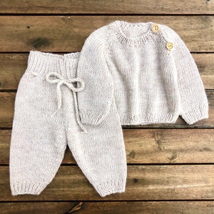 Op de afbeelding: Een gebreide baby-outfit in een licht beige kleur. De outfit bestaat uit een longsleeve trui met twee houten knopen en een bijpassende broek met een trekkoord.