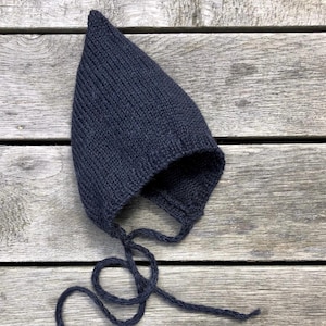 Peut inclure: Un bonnet de bébé en tricot noir avec un sommet pointu et des liens. Le bonnet est posé sur une surface en bois.