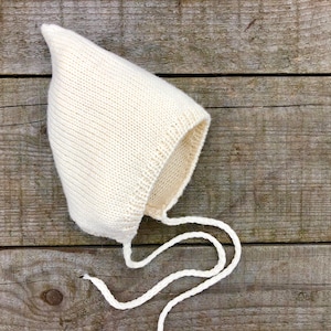 May include: A white knitted baby bonnet with a pointed top and a long tie. The bonnet is lying on a wooden surface.