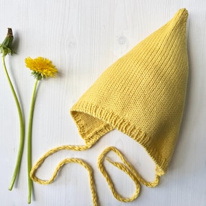 Puede incluir: Un gorro de bebé amarillo tejido con lazos largos, sobre una superficie blanca. El gorro tiene una parte superior puntiaguda y un borde acanalado. Una flor de diente de león amarilla y una cabeza de semilla están al lado del gorro.