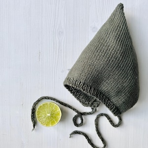 Può includere: Un berretto per neonato di lana verde con la cima a punta e lacci. Un spicchio di lime è in primo piano.